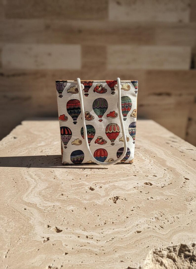May include: A small tote bag with a repeating pattern of colorful hot air balloons and clouds on a cream background. The bag has a rectangular shape with a white rope handle.