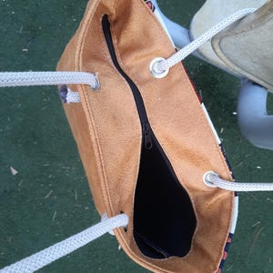 May include: A tan tote bag with a black zipper and white rope handles. The bag has a textured appearance and a patterned fabric detail at the top. The interior is black.