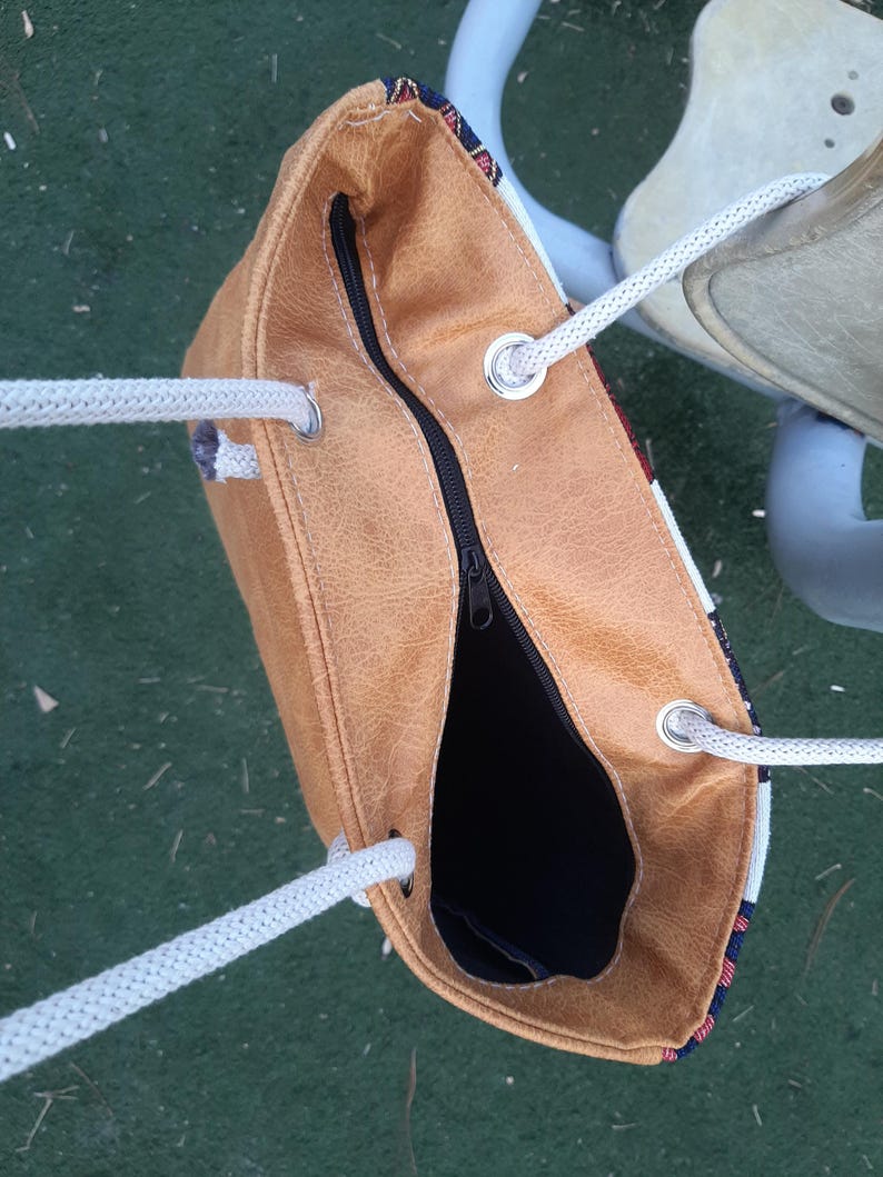 May include: A tan tote bag with a black zipper and white rope handles. The bag has a textured appearance and a patterned fabric detail at the top. The interior is black.