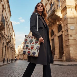 May include: A woman walking down a cobblestone street, wearing a dark gray trench coat and carrying a tote bag. The tote bag features a colorful hot air balloon pattern against a cream background.
