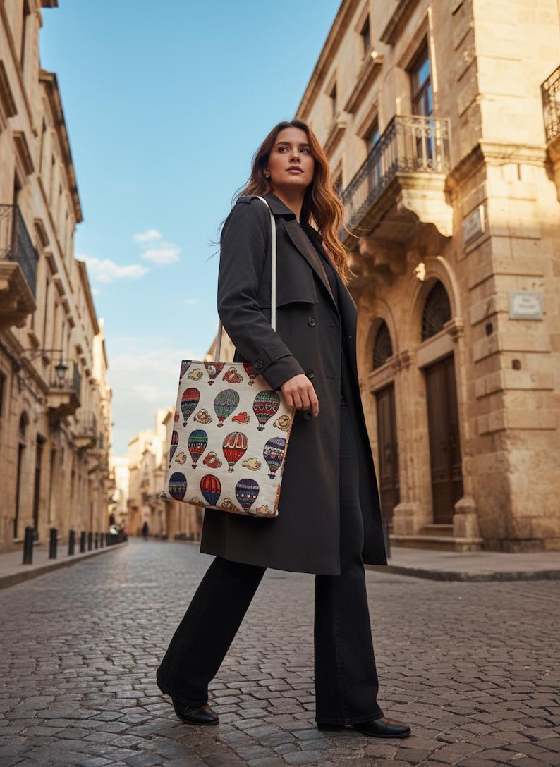 May include: A woman walking down a cobblestone street, wearing a dark gray trench coat and carrying a tote bag. The tote bag features a colorful hot air balloon pattern against a cream background.
