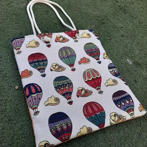 May include: A rectangular tote bag with a repeating pattern of colorful hot air balloons and clouds on a cream background. The bag has white handles and an orange trim. The balloons are red, blue, green, and purple.