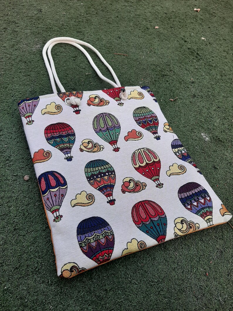 May include: A rectangular tote bag with a repeating pattern of colorful hot air balloons and clouds on a cream background. The bag has white handles and an orange trim. The balloons are red, blue, green, and purple.