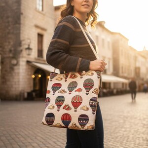 May include: A beige tote bag with a colorful hot air balloon pattern. The bag has a cream-colored strap and is being carried by a person walking on a cobblestone street.