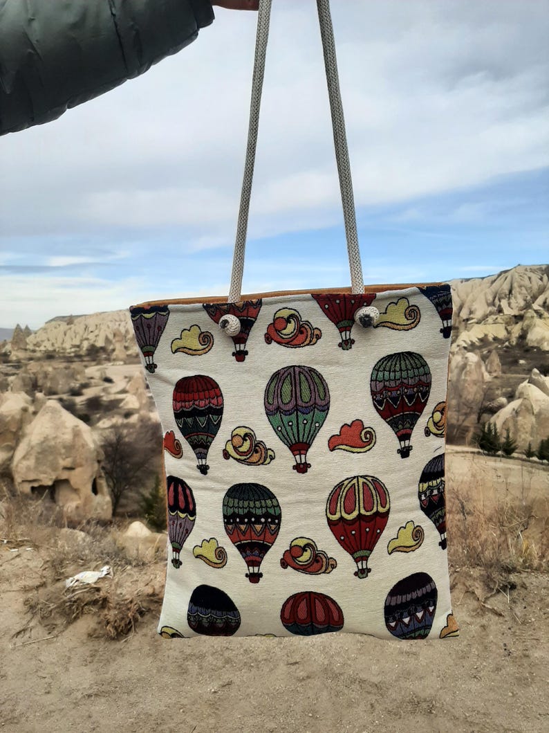 May include: A white tote bag with a repeating pattern of colorful hot air balloons and clouds. The bag has a light brown trim and gray rope handles. The background shows a landscape with rock formations and a cloudy sky.