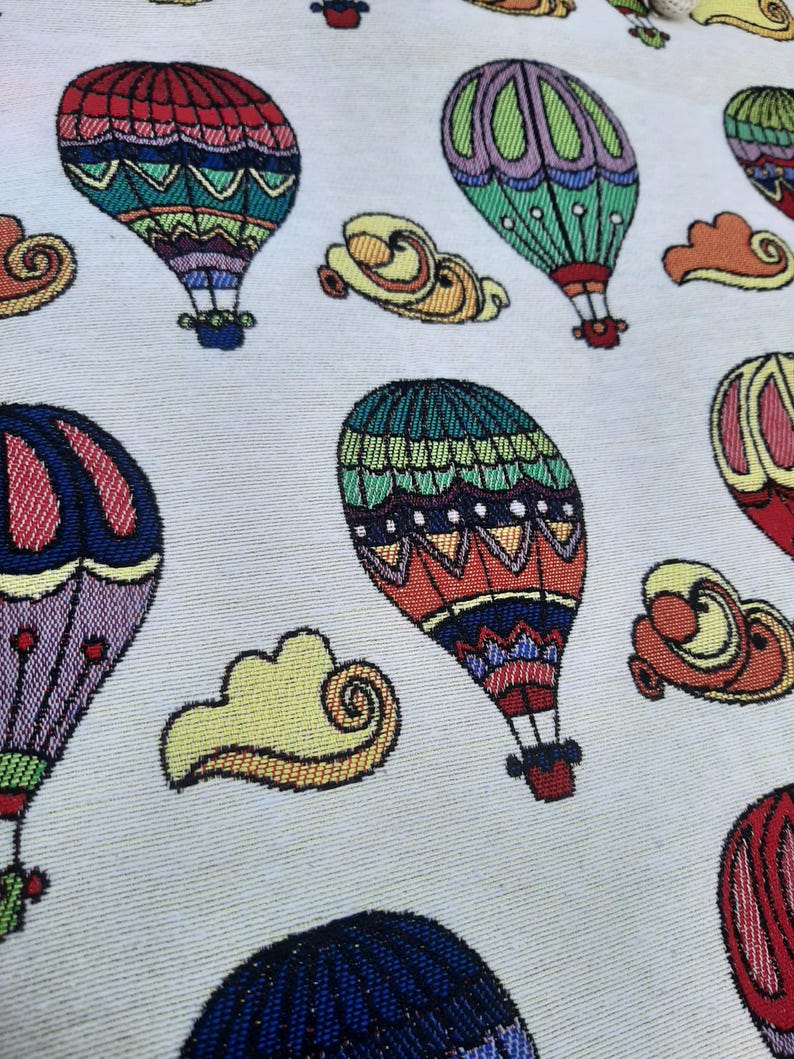 May include: A fabric with a repeating pattern of colorful hot air balloons and yellow cloud shapes on a light background. The balloons feature intricate designs in red, green, blue, and purple.