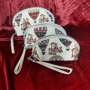 May include: Three off-white, half-moon shaped cosmetic bags with a hot air balloon design in red, brown, and beige. Each bag has a matching wrist strap and a silver zipper. The bags are arranged on a red velvet surface.
