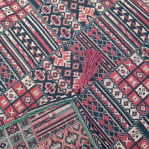 May include: A red, green, and black patterned throw blanket with a fringe tassel. The blanket has a geometric design with a variety of shapes and colors.