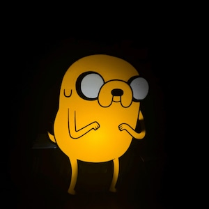 May include: A yellow cartoon character with large white eyes, a small nose, and a wide smile, set against a black background. The character is illuminated, creating a glowing effect. The character's arms are bent at the elbows.