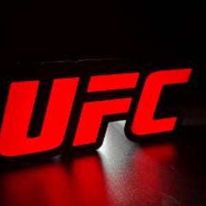 Puede incluir: Un logotipo de la UFC negro y rojo, iluminado sobre un fondo oscuro. Las letras "UFC" son en negrita y de color rojo vibrante, con un brillo sutil. El logotipo es probablemente un letrero o un emblema.