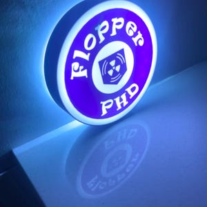 May include: A round, illuminated sign with a purple and white design. The sign features the text "Flopper PHD" in white, with a radiation symbol in the center. The sign is lit with a blue glow, creating a striking visual effect.