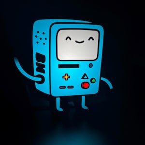 May include: A glowing blue cartoon character with a smiling face on a white screen. The character has arms, legs, and a control panel with buttons. The letters "BM" are visible on the side. The background is black.