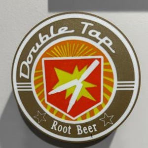Puede incluir: Un letrero redondo marrón y blanco de Double Tap Root Beer. El letrero presenta un emblema rojo y amarillo con un diseño de rayo blanco. Las palabras "Double Tap" y "Root Beer" están escritas en blanco.