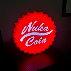 Peut inclure: Un panneau lumineux en forme de bouchon de bouteille Nuka Cola rouge. Le panneau présente le logo Nuka Cola en écriture blanche sur un fond rouge. Le panneau a un bord festonné et est éclairé de l'intérieur.