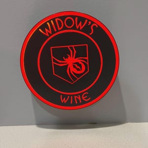 Puede incluir: Un posavasos redondo y negro con texto rojo iluminado y un emblema de araña roja. El texto dice "WIDOW'S WINE". El emblema de la araña está dentro de un escudo.