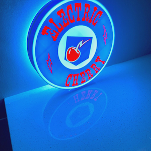 Puede incluir: Un letrero redondo e iluminado con las palabras "ELECTRIC CHERRY" en letras rojas. El letrero presenta un fondo azul con un gráfico central blanco y azul de una cereza. El letrero está iluminado con un brillo azul.