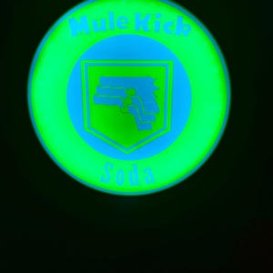 Puede incluir: Un letrero circular verde y azul brillante con las palabras "Mule Kick Soda". El letrero presenta un gráfico azul de un arma dentro de un escudo. El fondo es negro.