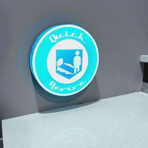 Peut inclure: Un panneau rond et lumineux avec une bordure turquoise et un texte blanc qui dit "Quick Revive". Le graphisme central représente une personne à côté d'un lit. Le panneau est monté sur un mur gris.