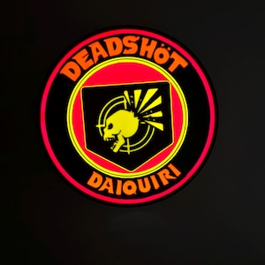 May include: A circular emblem with the words "DEADSHOT DAIQUIRI" in orange lettering. The emblem features a black background with a red and yellow border. The center has a yellow skull and sunburst design.