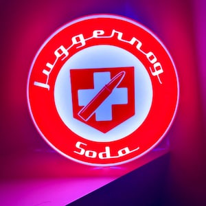 May include: A round, illuminated sign with a red border and a white interior. The sign features a red shield with a white cross and a bullet, and the word "Juggernog" in white script. The word "Soda" is also written in white script.