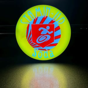 May include: A neon yellow disc with the text "Jamin-Up Joda" in blue. The center features a red shield with a blue and white design of a person running. The disc is illuminated against a dark background.