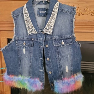 May include: A blue denim vest with a white lace collar and rainbow faux fur trim. The vest has a button closure and two pockets. The label on the vest reads "a series".