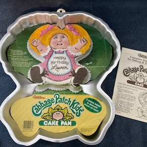 May include: A silver metal cake pan shaped like a Cabbage Patch Kid doll. The pan is designed to bake a cake in the shape of a doll. The pan includes instructions for baking and decorating the cake. The instructions include a picture of a doll cake with the text "Happy Birthday Lauren".
