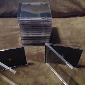 May include: A stack of clear, empty CD jewel cases, with a few cases open and lying in front. The cases are made of transparent plastic with a black interior. The background is a brown, textured surface.