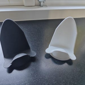 Op de afbeelding: Twee oorvormige plastic objecten. Eén is zwart, de andere wit. Beide hebben een afgeronde bovenkant en een uitlopende basis met twee kleine, gebogen verlengingen. Ze liggen op een donker, getextureerd oppervlak.