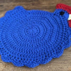 May include: A handmade, blue crochet trivet in the shape of a rooster. The trivet features a red comb and wattle, and a black eye. The trivet is circular with a scalloped edge, and is designed to protect surfaces from hot cookware.