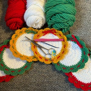 May include: A collection of handmade crochet coasters in red, white, and green, with yellow and green scalloped edges. Yarn balls in matching colors and crochet tools are also visible.