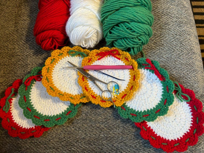 May include: A collection of handmade crochet coasters in red, white, and green, with yellow and green scalloped edges. Yarn balls in matching colors and crochet tools are also visible.