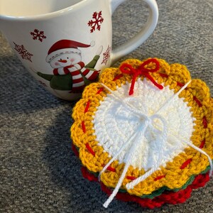 May include: A white ceramic mug with a snowman design and red snowflake accents. A stack of crocheted coasters in red, green, yellow, and white, tied with white string and a red bow, sits beside the mug.