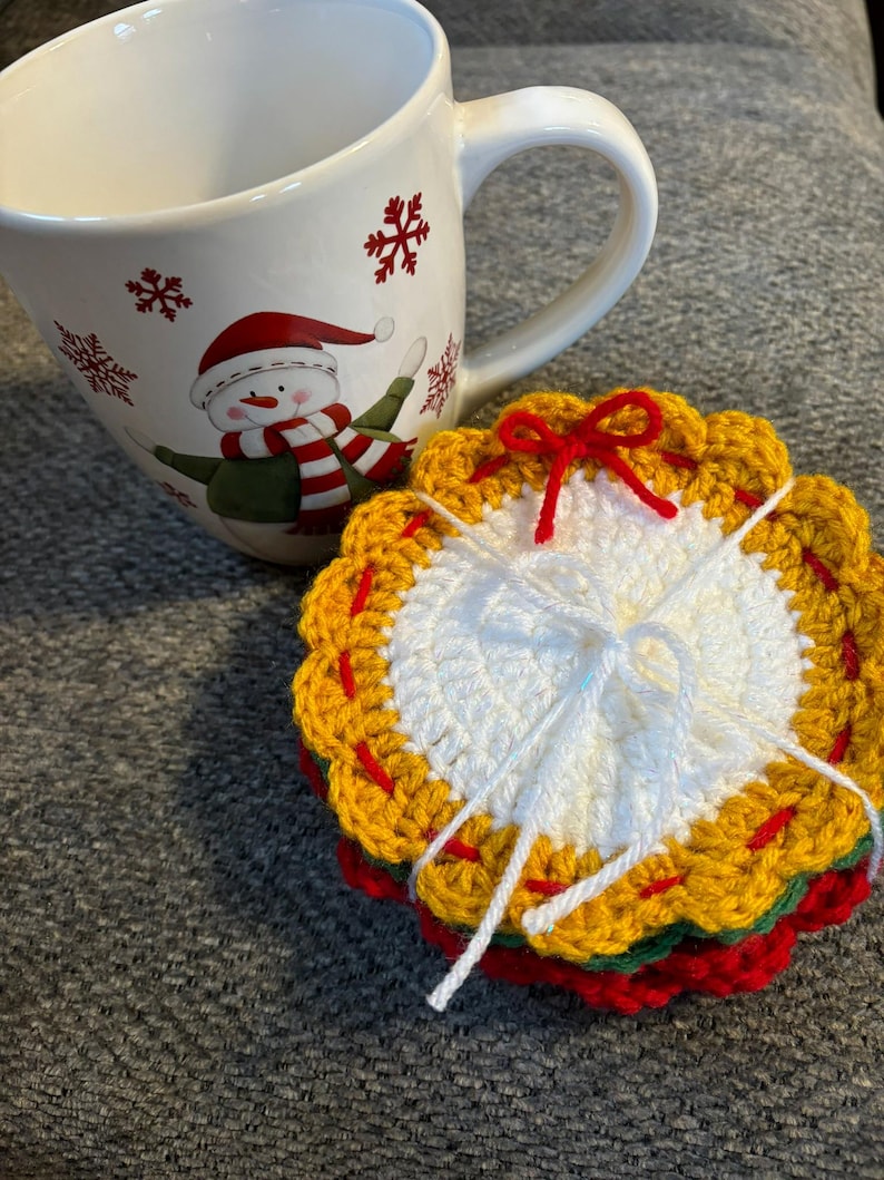 May include: A white ceramic mug with a snowman design and red snowflake accents. A stack of crocheted coasters in red, green, yellow, and white, tied with white string and a red bow, sits beside the mug.