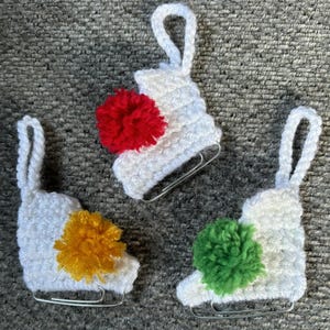 Könnte beinhalten: Drei weiße gehäkelte Schlittschuh-Ornamente mit roten, gelben und grünen Bommeln.