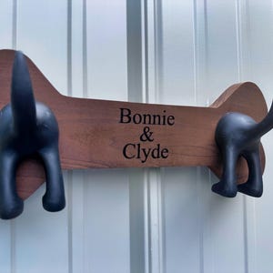 May include: A wooden dog bone-shaped wall hook with two black dog butt hooks. The text "Bonnie & Clyde" is printed in black on the brown wood. The background is a white wall.