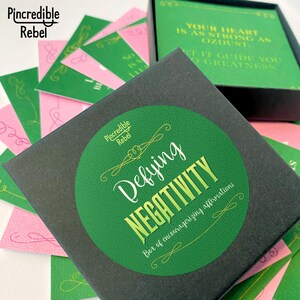 May include: A black box with a green circle sticker that says "Defying Negativity" and "Box of encouraging affirmations". The box is filled with pink and green cards.