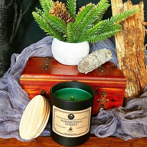 May include: A black candle with a wooden lid and a label that reads "Foreboding Forest." The candle has a green wax and is set on a wooden chest with a white vase of greenery and a bundle of sage. A rustic, nature-themed aesthetic.