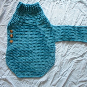 Op de afbeelding: Een turquoise gehaakte trui met een hoge kraag en &eacute;&eacute;n lange mouw. De trui heeft drie bruine knopen aan de voorkant. Het handgemaakte kledingstuk wordt op een wit oppervlak getoond.