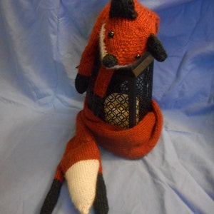 May include: A red and black knitted fox scarf with a fox head at the top. The scarf is wrapped around a black metal object.