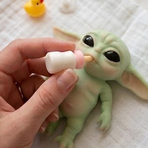 May include: A light green creature figurine with large black eyes, being fed with a small white and pink toy bottle. A tiny yellow rubber duck and a white pacifier are positioned nearby on a white textured fabric surface.