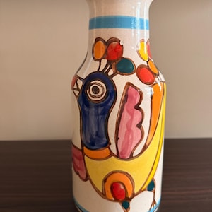 May include: A hand-painted ceramic vase with a colorful bird design. The vase features a cream-colored base with a blue stripe, and a brown rim. The bird is depicted with yellow, blue, pink, orange, and red accents.
