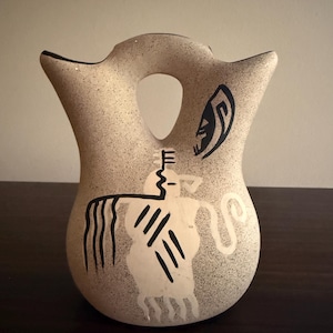 May include: A beige ceramic wedding vase with a speckled texture and two spouts connected by a central handle. The front features a black and white painted graphic of a stylized figure with long flowing lines and a separate crescent-shaped motif.