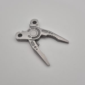 Könnte beinhalten: Ein silberfarbenes Leatherman Multitool-Zangenkopf-Bauteil aus Edelstahl mit einem zentralen Drehpunkt, gezahnten Greifbacken und zwei separaten Befestigungsarmen mit kreisförmigen Aussparungen sowie einer durchgehend gebürsteten Metalloberfläche.