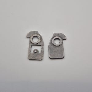 May include: Two small, gray metal brackets with a rectangular shape and a circular hole. One bracket has a curved top and a small rectangular cutout. The other bracket is a simple rectangle with a circular hole.