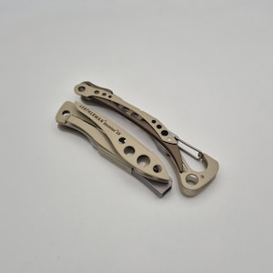 Könnte beinhalten: Ein zusammengeklapptes Leatherman Skeletool CX Multifunktionswerkzeug mit metallisch beigem und dunkelgrauem Finish. Das Werkzeug verfügt über einen skelettierten Rahmen mit kreisförmigen Aussparungen, einen Taschenclip und einen Karabiner.