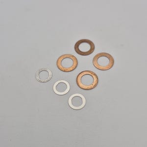 May include: A collection of seven metal washers, four are copper coloured and three are silver coloured. The washers are arranged in a semi-circle pattern on a white surface.