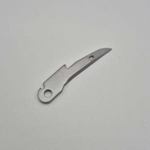 May include: A small, silver-colored multi-tool with a bottle opener, a small blade, and a hole for attaching to a keychain. The tool is made of metal and has a simple, utilitarian design.