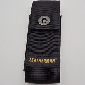 May include: Black nylon Leatherman sheath with a snap closure. The sheath has a rectangular shape with a flap that secures with a metal snap. The Leatherman logo is printed in gold on the front.