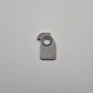 May include: A small, gray metal component with a circular hole and a curved top edge. The piece has a rectangular base with a rounded corner and a textured surface. The object is set against a plain, light background.